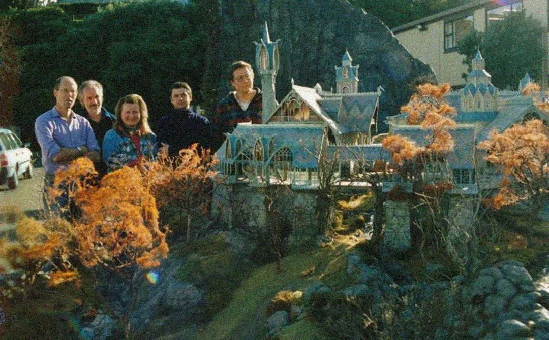 A group of five people stand outdoors beside an elaborate miniature model of a fantasy castle with detailed buildings, towers, and orange autumn trees.