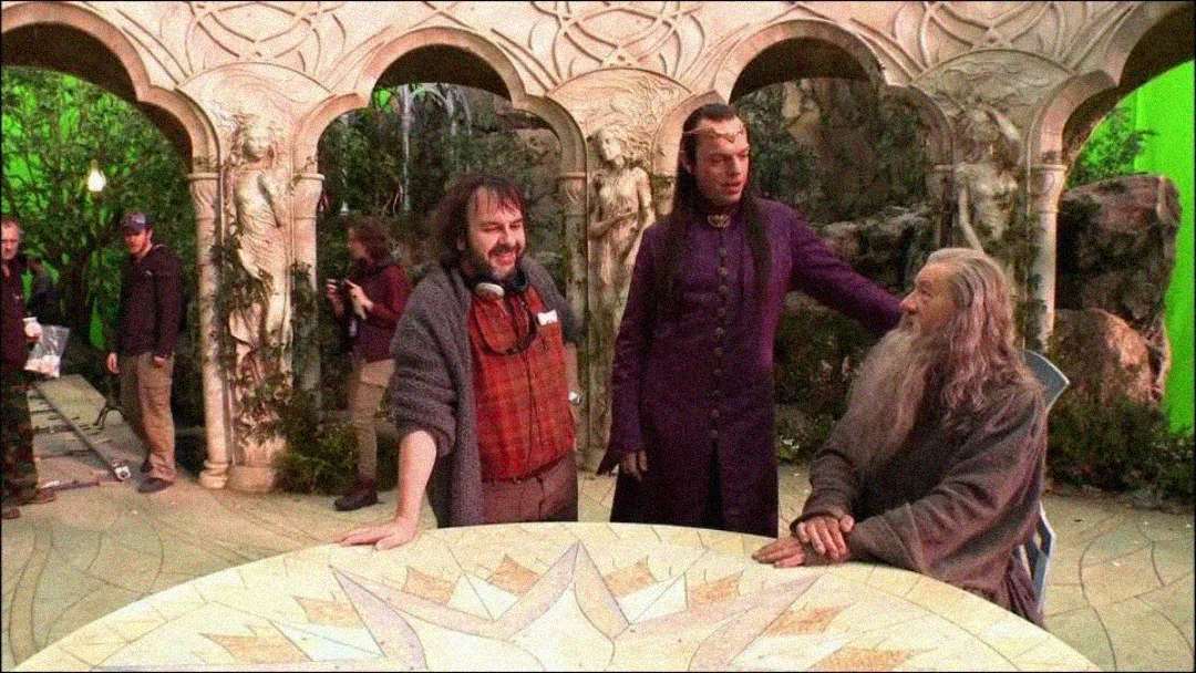 Three men, including one in a burgundy robe and another with long gray hair and beard, stand and sit around a round, carved table on a detailed fantasy film set, with crew and green screens visible in the background.
