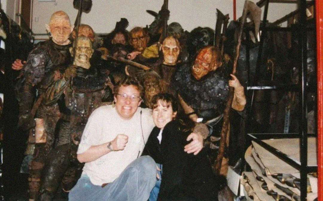 Two people crouch and smile for the camera, surrounded by actors in detailed orc costumes with weapons, in what appears to be a backstage or studio setting. The background is filled with props and equipment.
