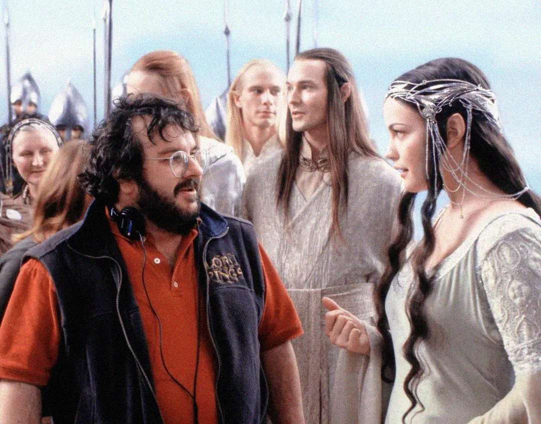 A man in modern clothes and headset stands with actors in elven costumes on a film set. One woman in a silver dress and headpiece speaks with him, while others in white robes stand behind them.
