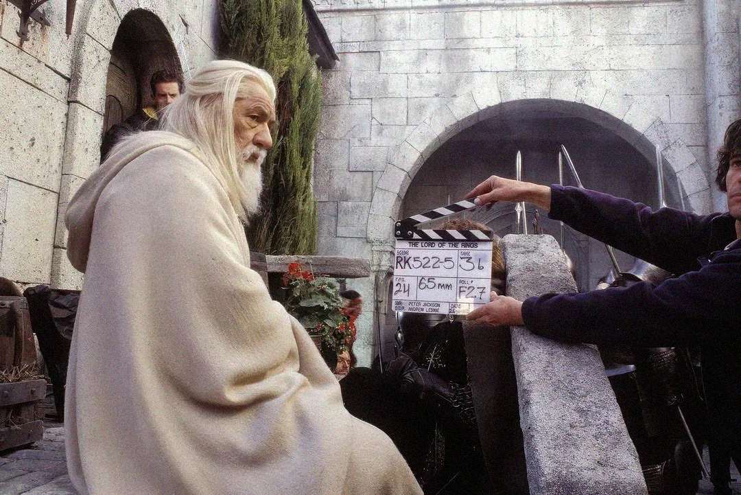 An actor dressed as Gandalf in a white robe sits on set, while a crew member holds a movie clapperboard labeled "Lord of the Rings" in front of him; medieval stone walls and props are visible in the background.