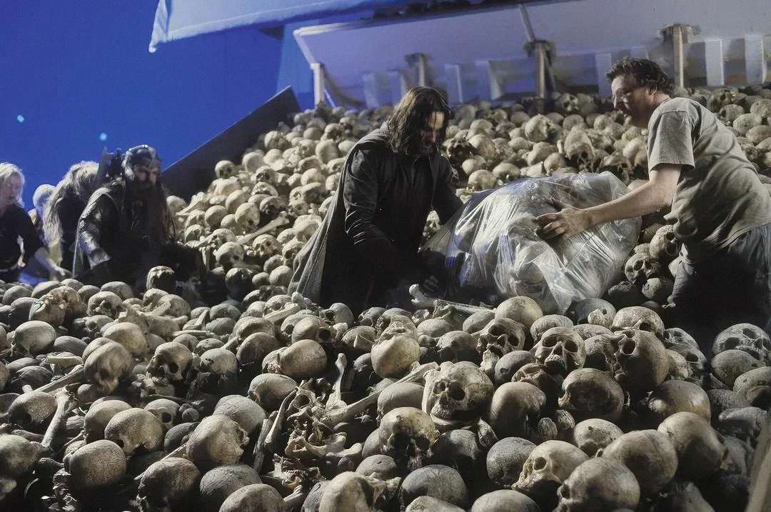 Actors and crew members work on a movie set filled with fake human skulls, with a blue screen in the background and film equipment visible.