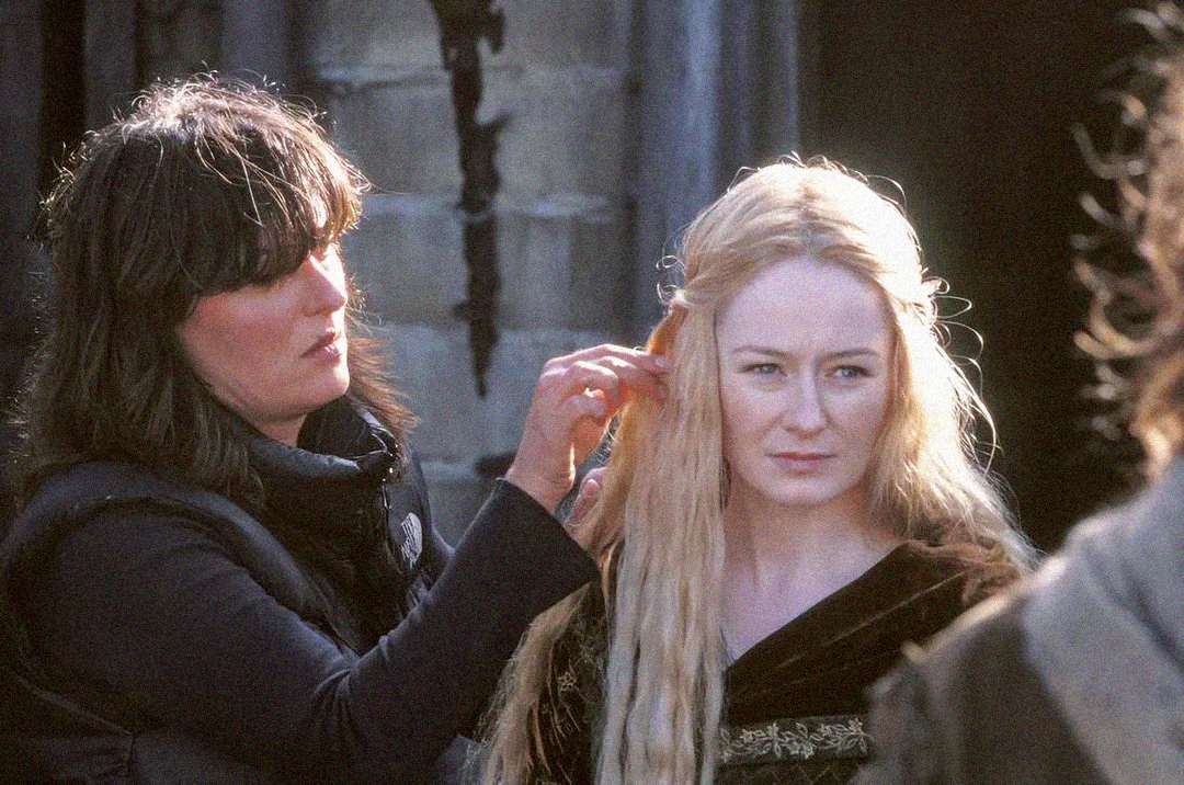 A crew member adjusts the hair of an actor in costume with long blond hair and medieval-style clothing, on a film set with stone walls in the background.