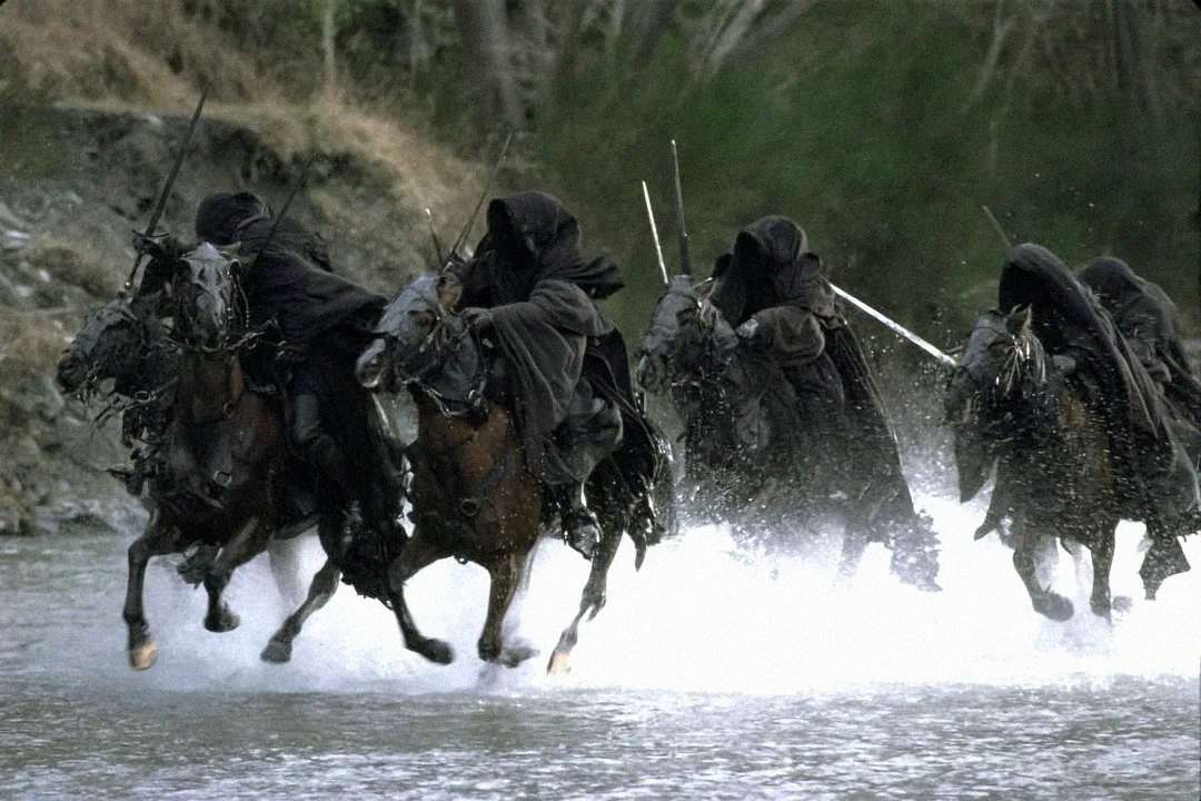 Four cloaked riders on horseback, dressed in black hooded robes, charge through a shallow, splashing river with a forested background. The scene is dramatic and conveys a sense of urgency and menace.
