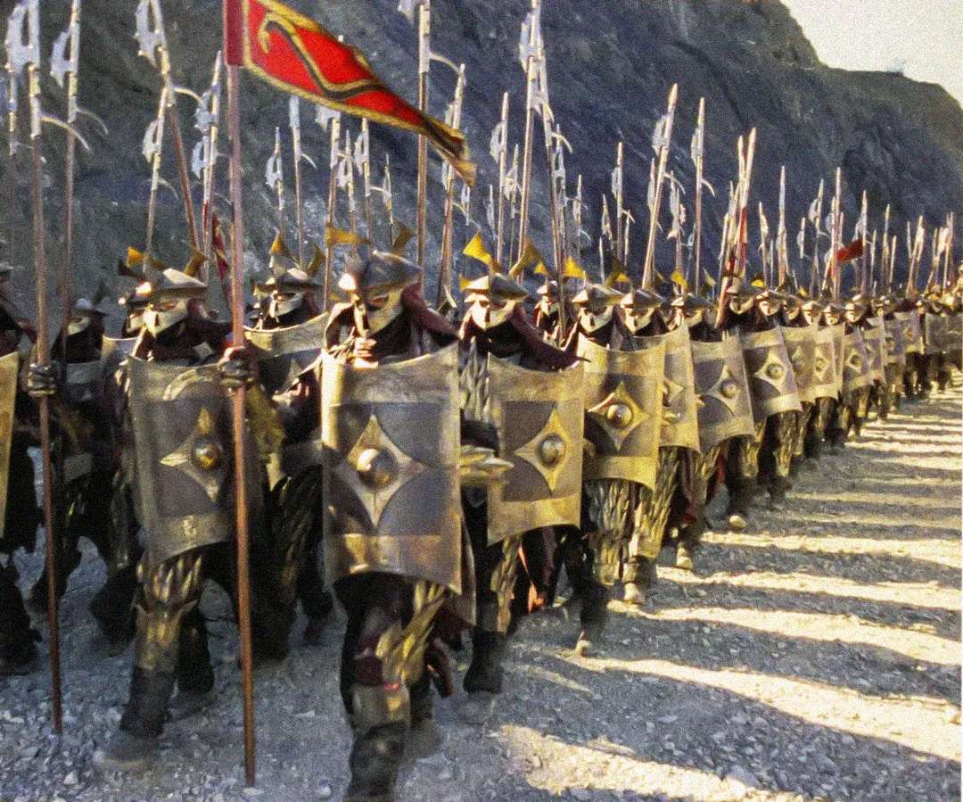 A large group of armored soldiers carrying spears and ornate shields march in formation along a rocky path, with a red flag leading the procession.