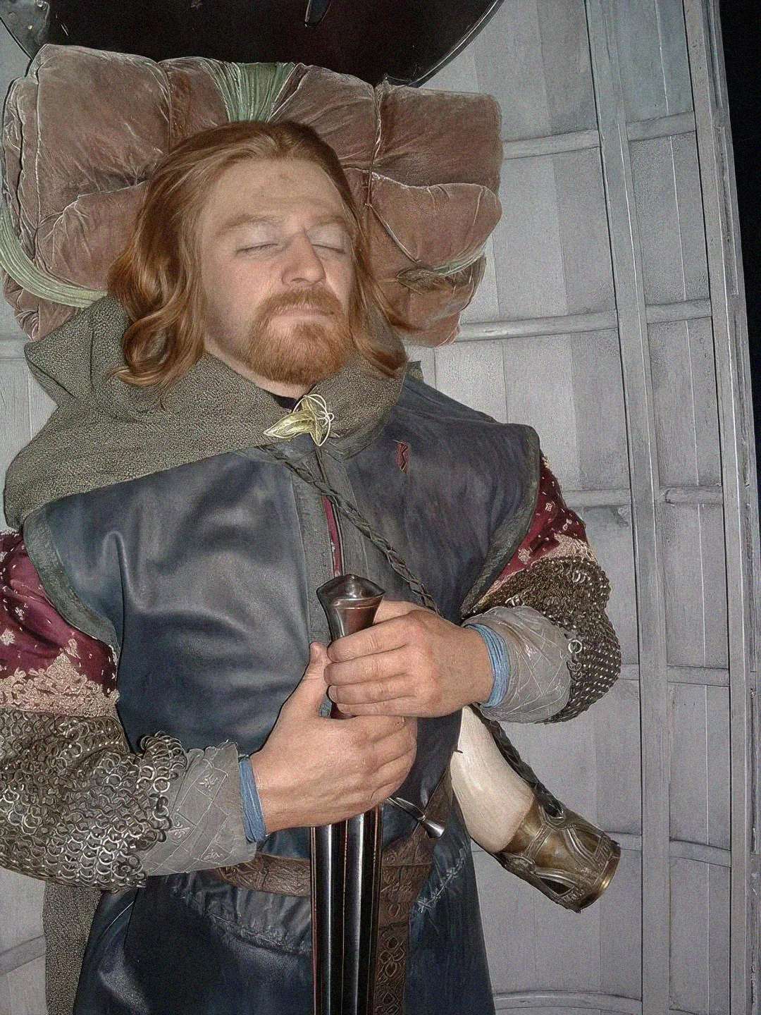 A man with long hair and a beard lies in a boat with his eyes closed, dressed in medieval-style clothing, holding a sword on his chest, with a horn at his side and a blanket under his head.