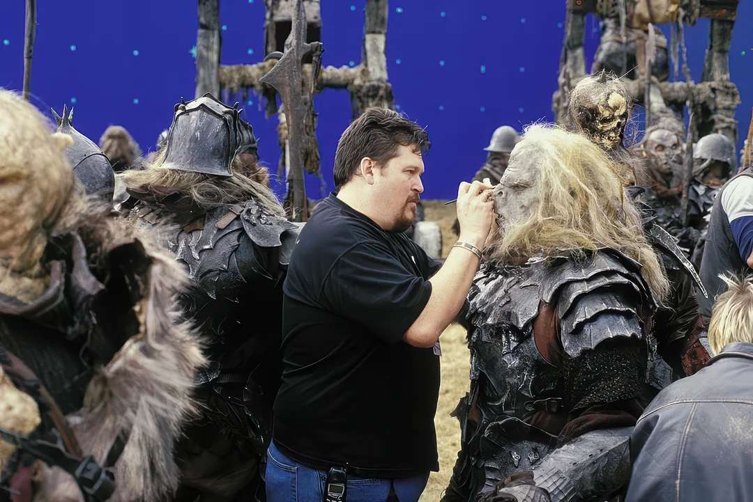 A makeup artist applies special effects makeup to an actor dressed as an orc on a movie set, surrounded by other actors in costume, with a blue screen background behind them.
