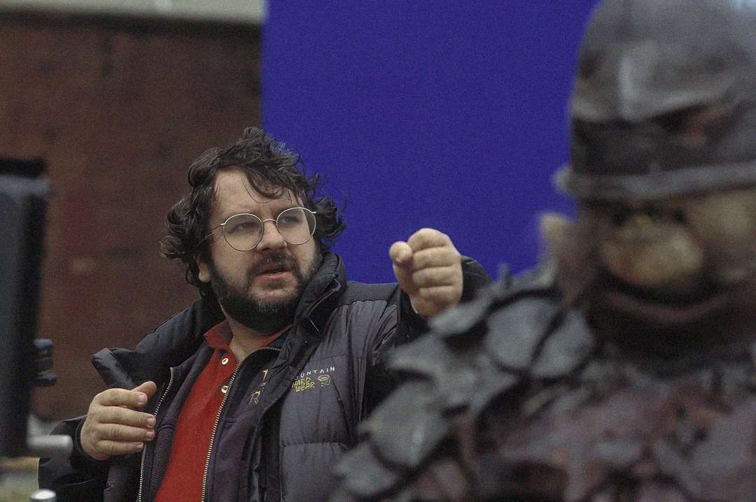 A man with glasses and a beard gestures with his hand while standing next to a person in a detailed monster costume with armor. The background is partially blue and blurred.