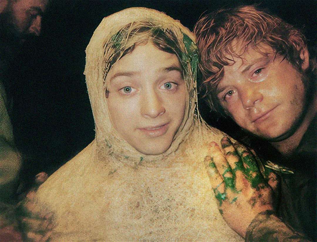 Two young men pose closely together. One is covered head to toe in fake spider webs and has leaves in his hair, while the other, with wet hair, smiles slightly. Both have makeup effects and appear to be on a film set.