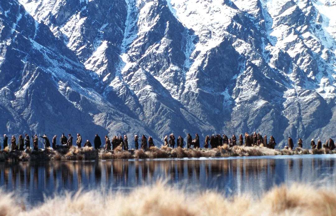 A line of people in dark cloaks walks along the edge of a calm lake, with tall snow-capped mountains rising steeply in the background. Golden grass lines the foreground.