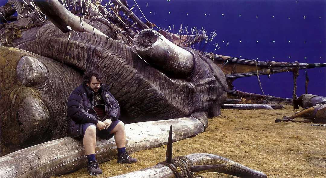 A person in a jacket and shorts sits on a large, fallen creature prop on a film set, with a blue screen background and scattered debris, suggesting a behind-the-scenes moment from a fantasy or adventure movie production.