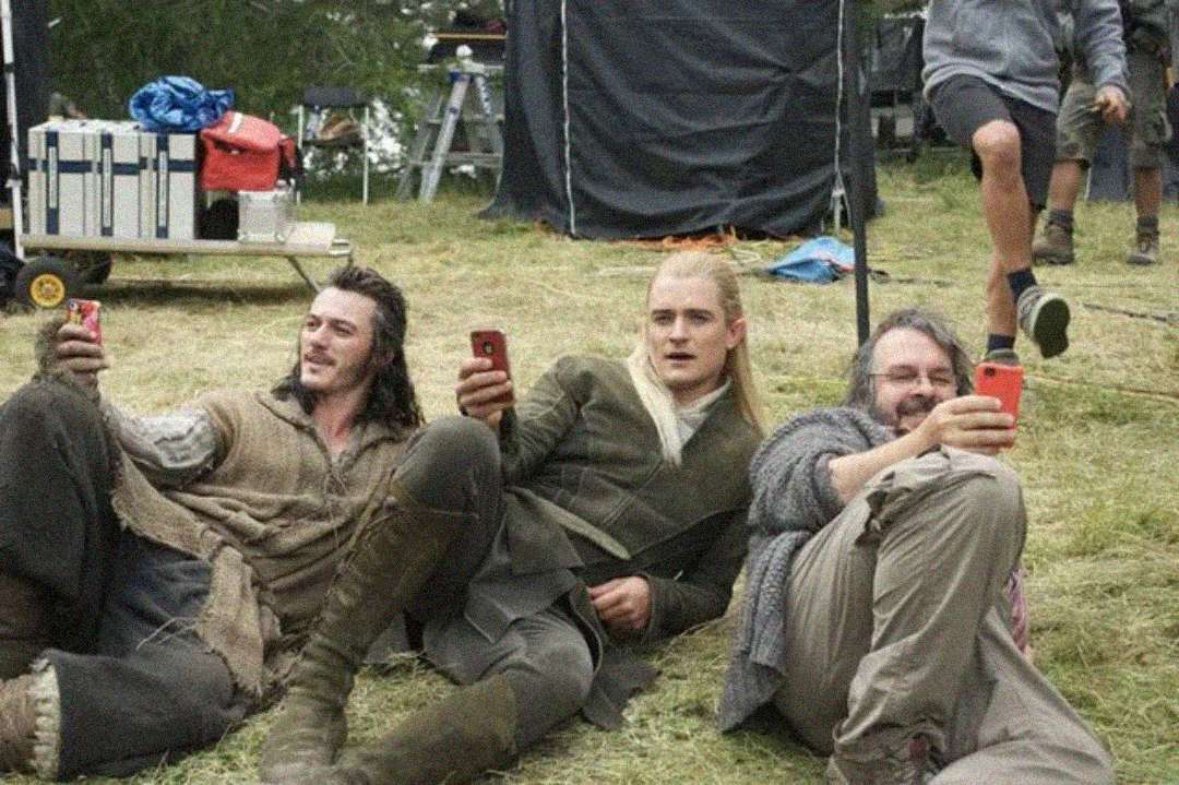 Three men in medieval-style costumes relax on grass, smiling and holding up red smartphones as if taking selfies. Equipment and crew are visible in the background, suggesting a film set.