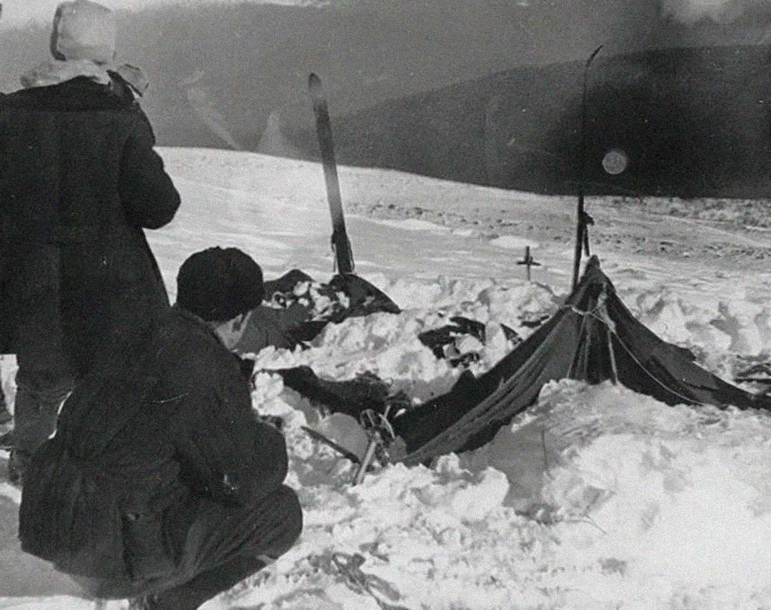 Two people in winter clothing examine a partially collapsed tent covered in snow on a snowy mountainside, with skis and a cross marking the site. The scene appears cold and desolate.