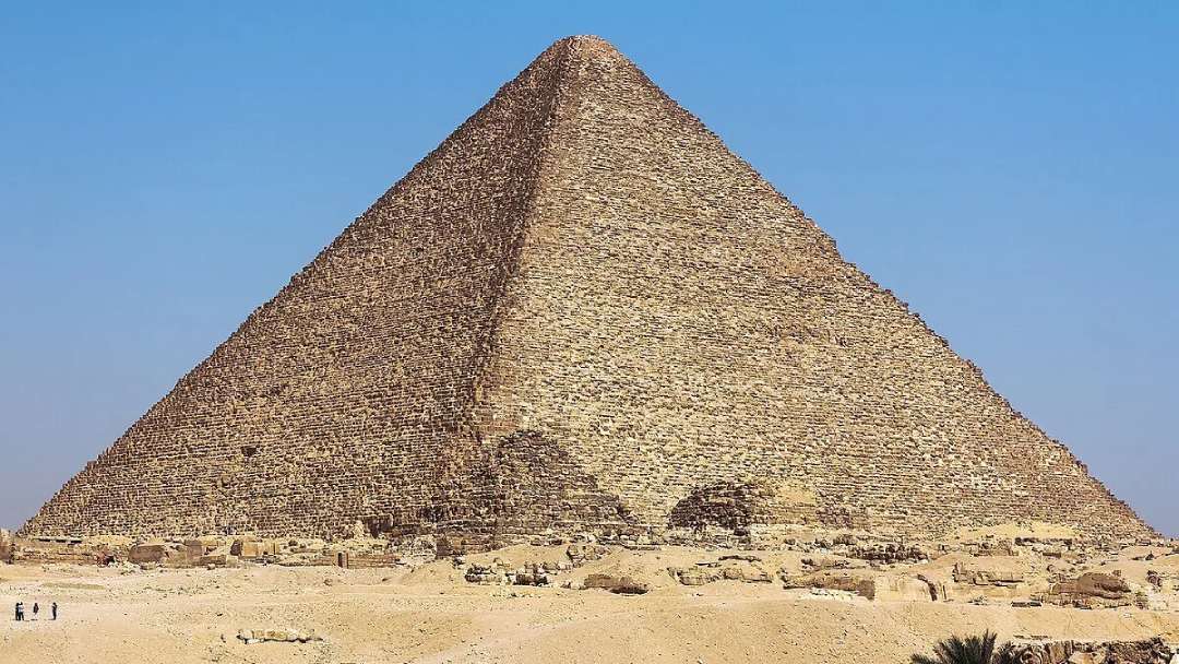 The Great Pyramid of Giza stands under a clear blue sky, its weathered limestone blocks forming a steep, angular structure. Small figures are visible at the base, highlighting the pyramid’s massive scale.