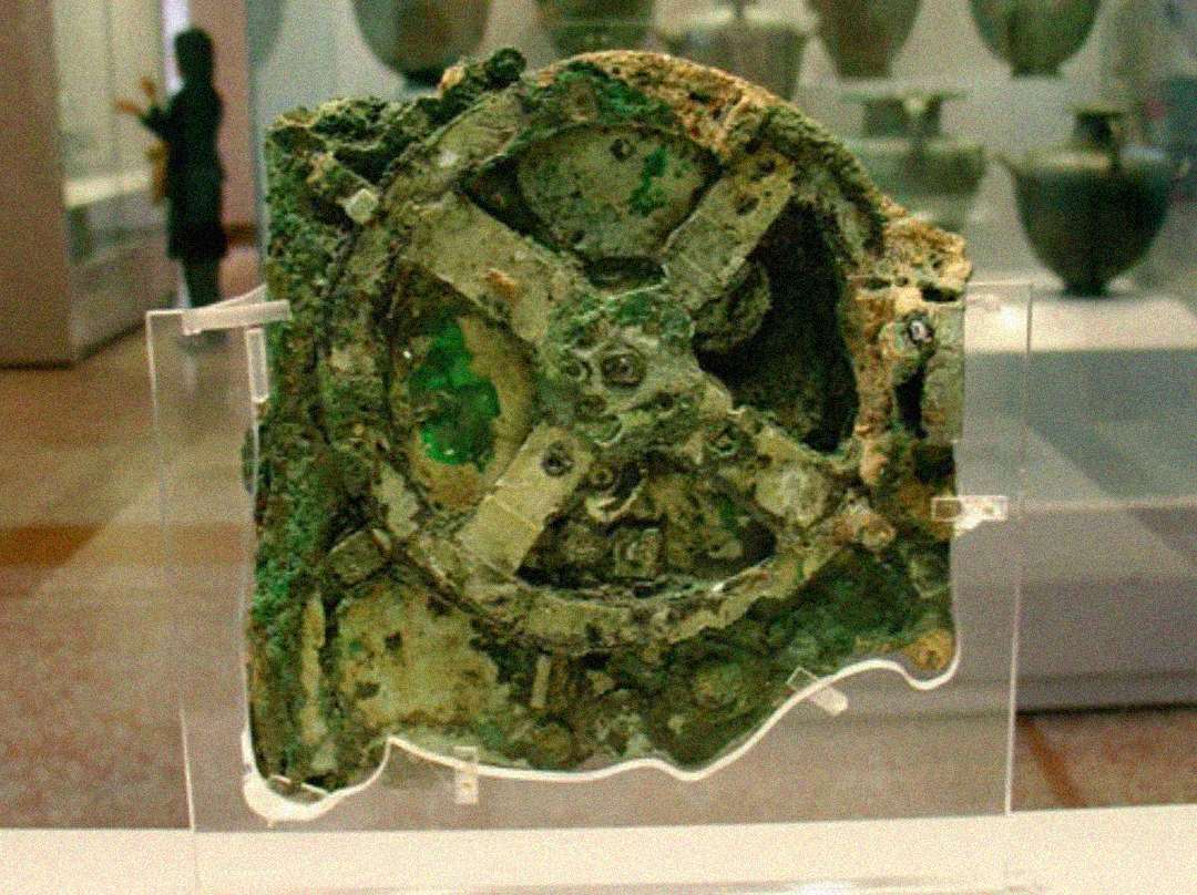 A corroded, ancient gear mechanism known as the Antikythera mechanism is displayed in a museum case, with shelves of artifacts and a blurred figure in the background.
