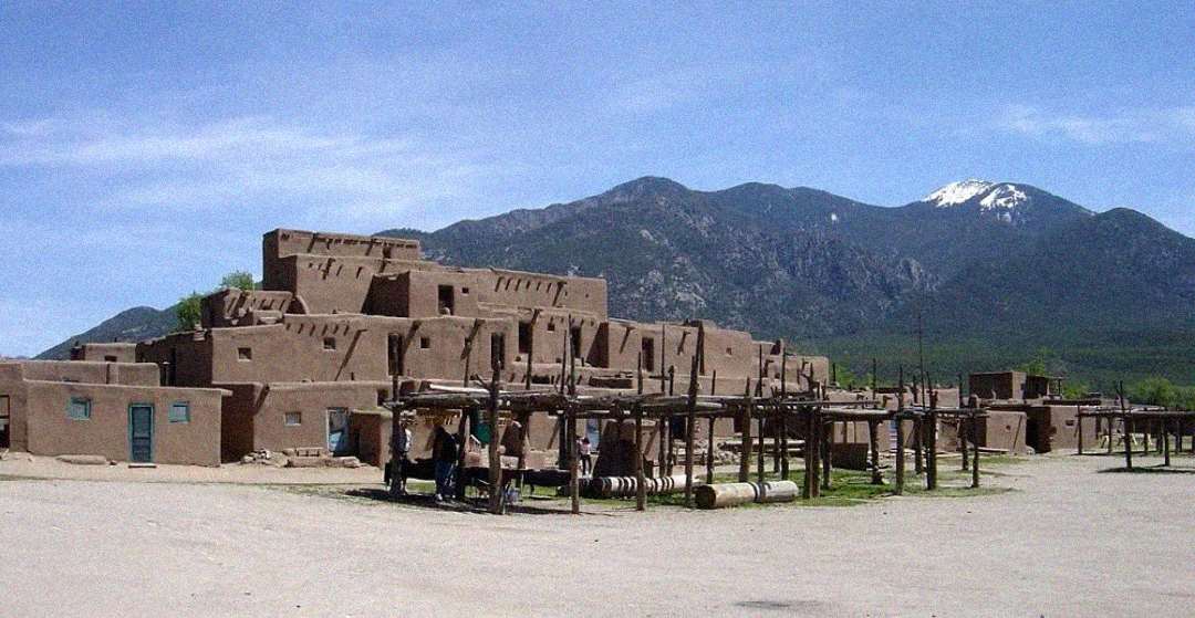 A large, multi-storied adobe pueblo building stands under a clear blue sky, surrounded by wooden structures, with mountains in the background and a few people gathered in the foreground.