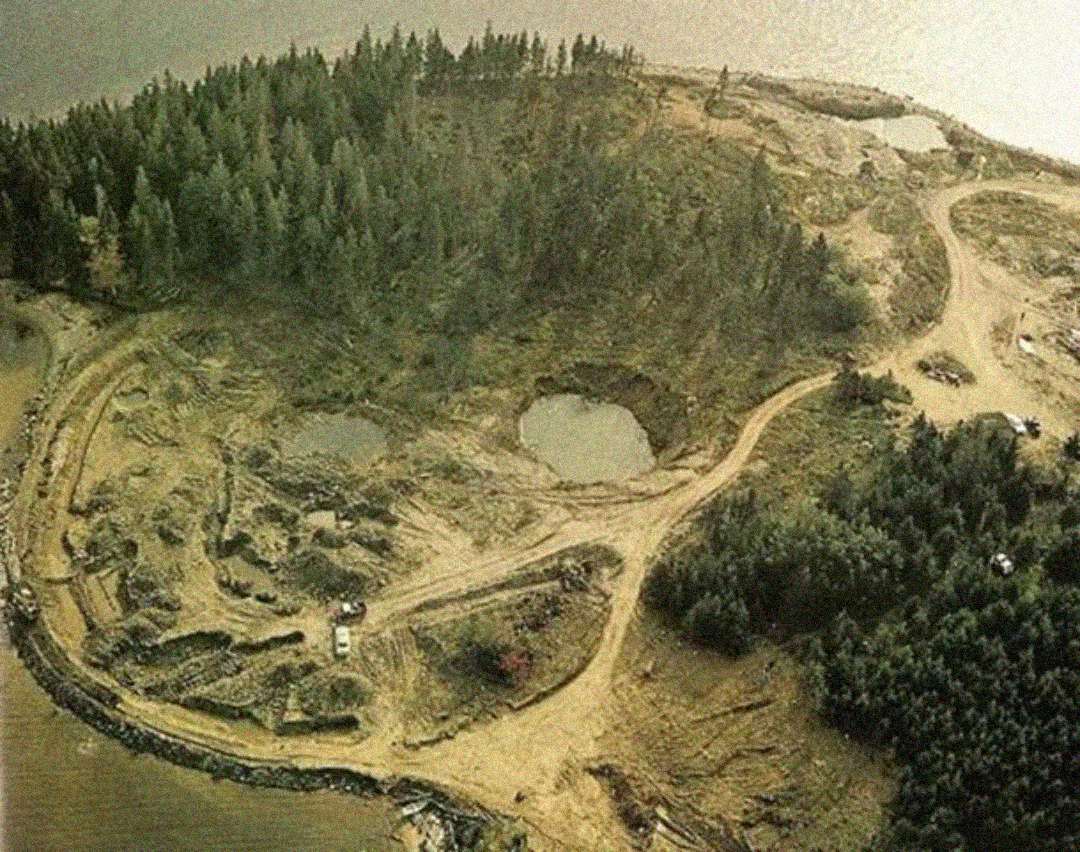Aerial view of a small island with dense trees, dirt roads, several large pits filled with water, and cleared areas indicating excavation or land alteration near the shoreline.