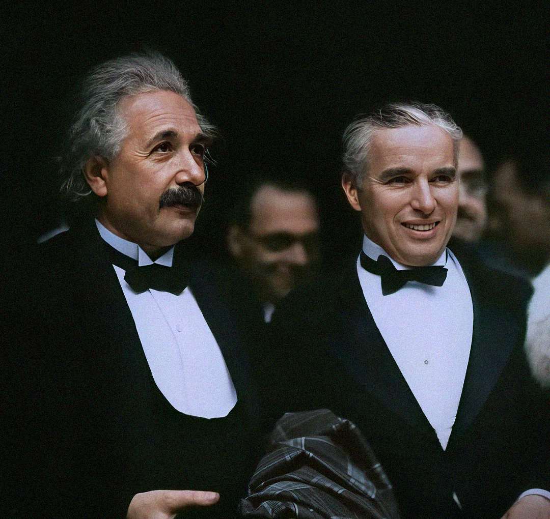 Two men in formal black tuxedos stand together, smiling. One has white hair and a mustache; the other has slicked-back hair. They appear to be at a formal event, with a dark background and other people partially visible behind them.