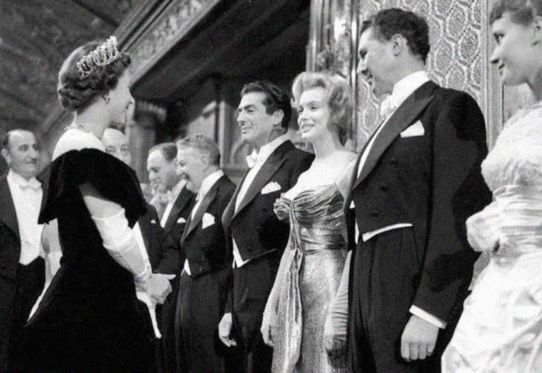 A black-and-white photo shows a group of elegantly dressed people in evening wear greeting a woman in a tiara and gown at a formal event, with ornate decor visible in the background.