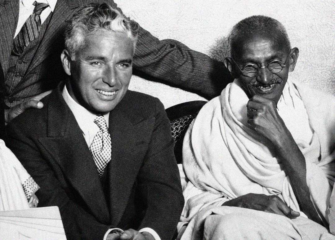 Two men are seated side by side, smiling. The man on the left wears a suit and tie, while the man on the right wears glasses and traditional Indian attire, with a shawl draped over his shoulder. Both appear relaxed and friendly.
