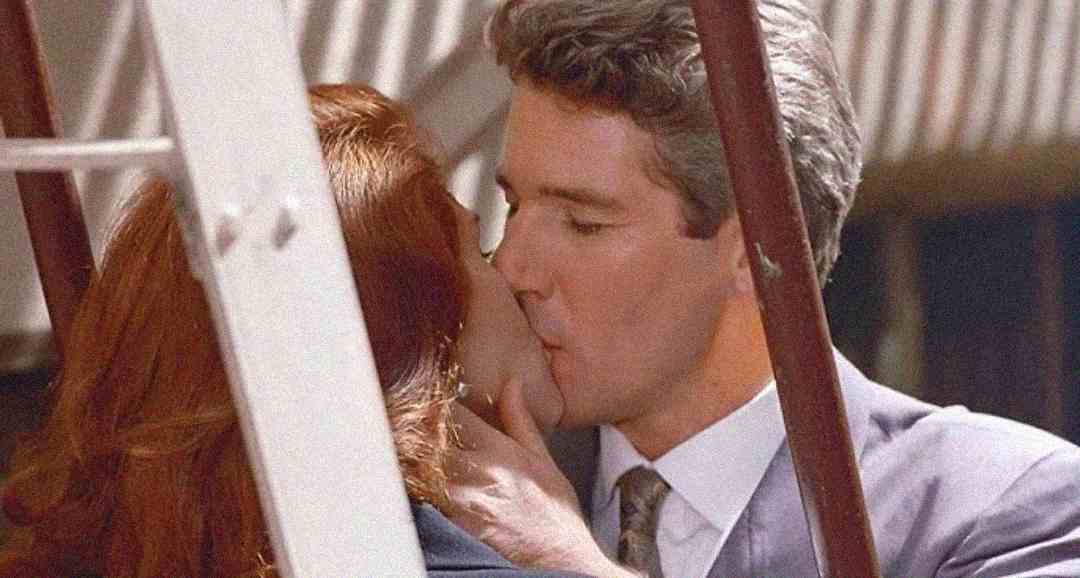 A man in a suit tenderly kisses a woman with red hair. They are framed by white metal bars, suggesting an intimate moment captured in a public or outdoor setting.