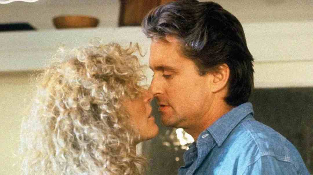 A man and woman stand very close to each other, appearing about to kiss. The woman has curly blonde hair and the man has dark hair and is wearing a blue button-up shirt. They are in an indoor setting.