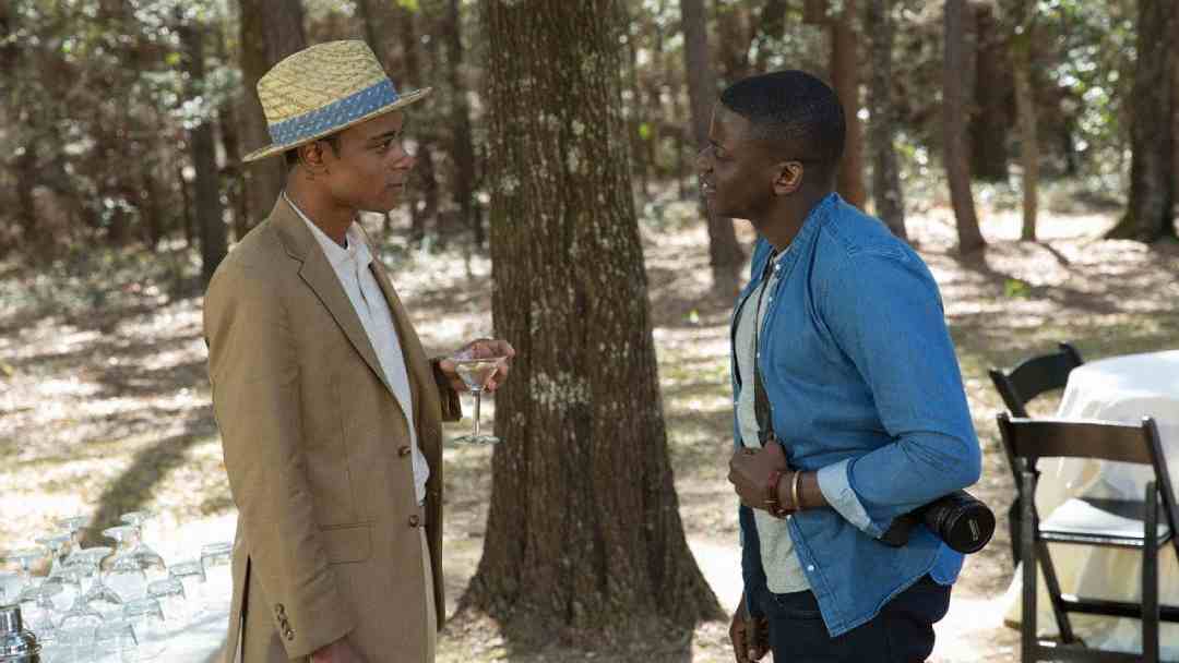 Two men stand and talk in a wooded outdoor setting. One wears a straw hat, beige jacket, and holds a drink; the other wears a blue shirt with a camera around his neck. A table with glasses and chairs is nearby.