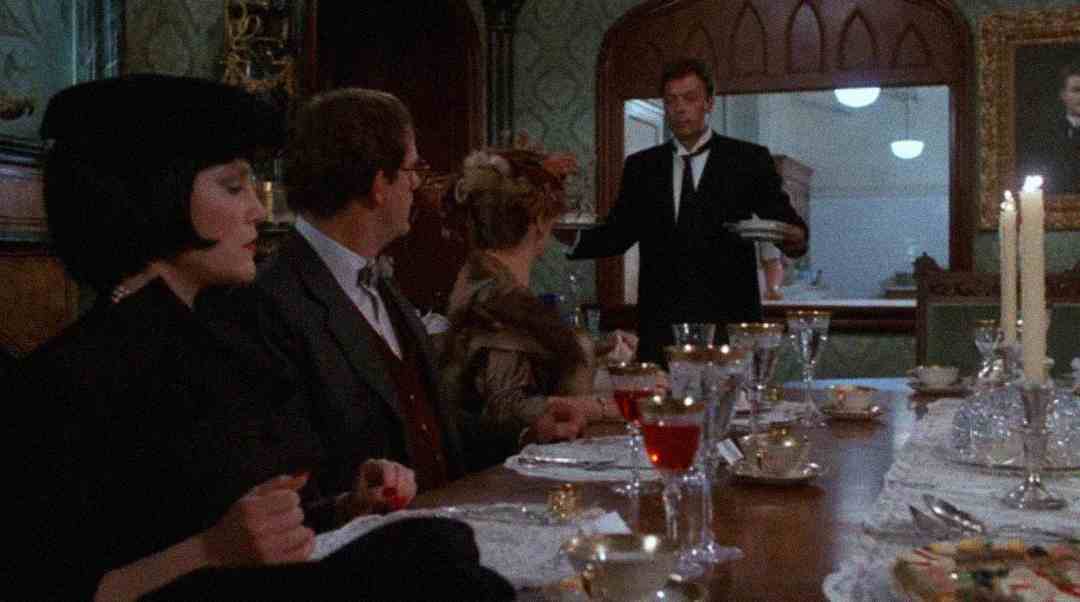Four people sit at an elegantly set dining table, with a man in a dark suit serving dishes. The room is dimly lit, featuring candles, fine china, and ornate decor. The atmosphere appears formal and tense.