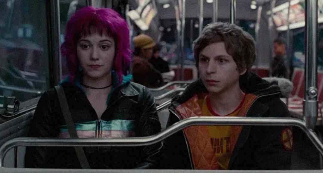 Two young people sit side by side on a bus at night. The woman has bright magenta hair and wears a black jacket, while the man has short brown hair and an orange jacket. Both look ahead with neutral expressions.