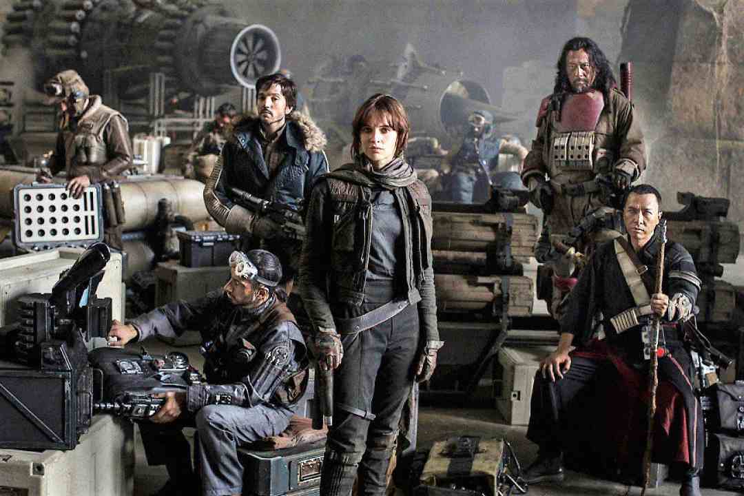 A group of five rebels in rugged clothing stand and sit among machinery and crates in a dimly lit, industrial setting, looking determined and ready for action.
