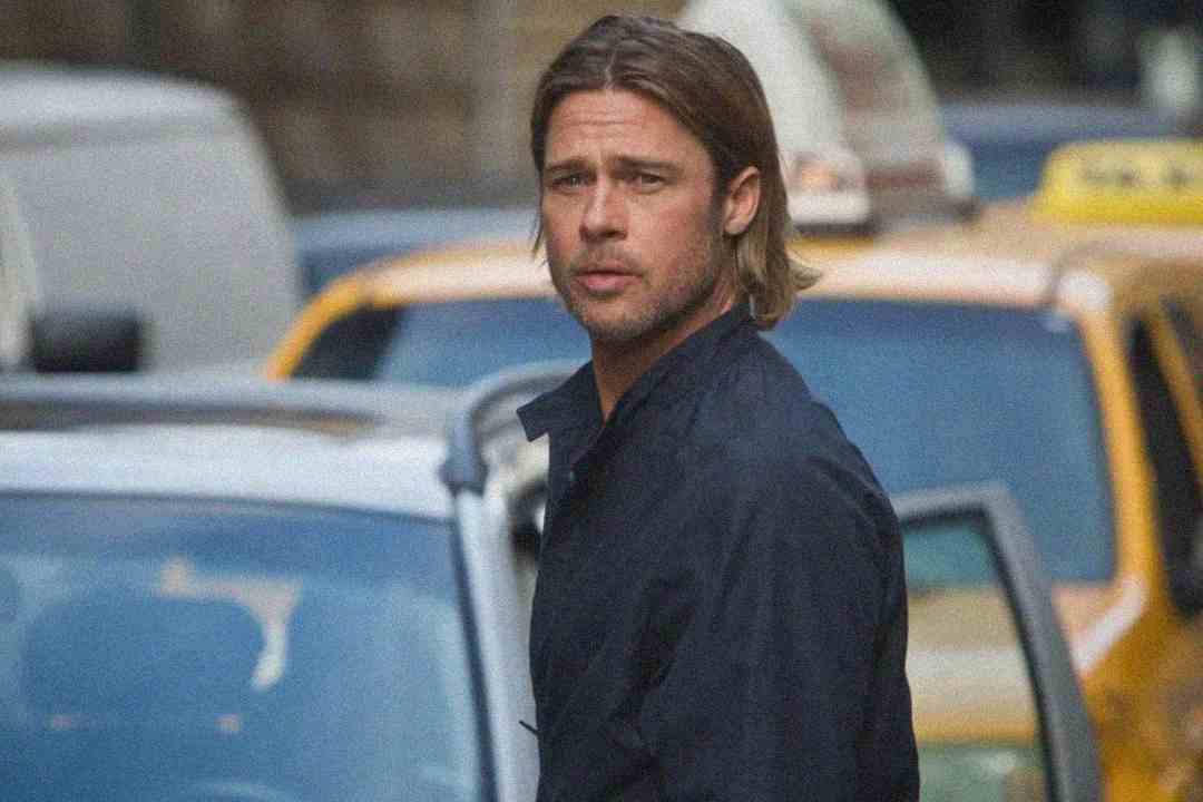 A man with shoulder-length hair and a dark jacket stands on a city street in front of cars, including a yellow taxi, looking over his shoulder with a serious expression.