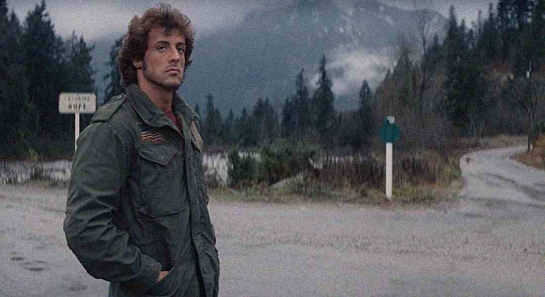 A man with curly hair wearing a green military jacket stands on a deserted road with a forest and mountains in the background under a cloudy sky.