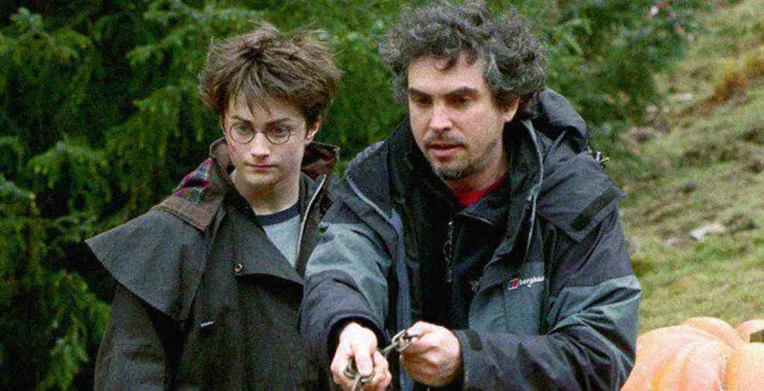 A person dressed as Harry Potter stands outdoors beside another person in a jacket, who appears to be giving instructions or demonstrating something. Trees and greenery are visible in the background.