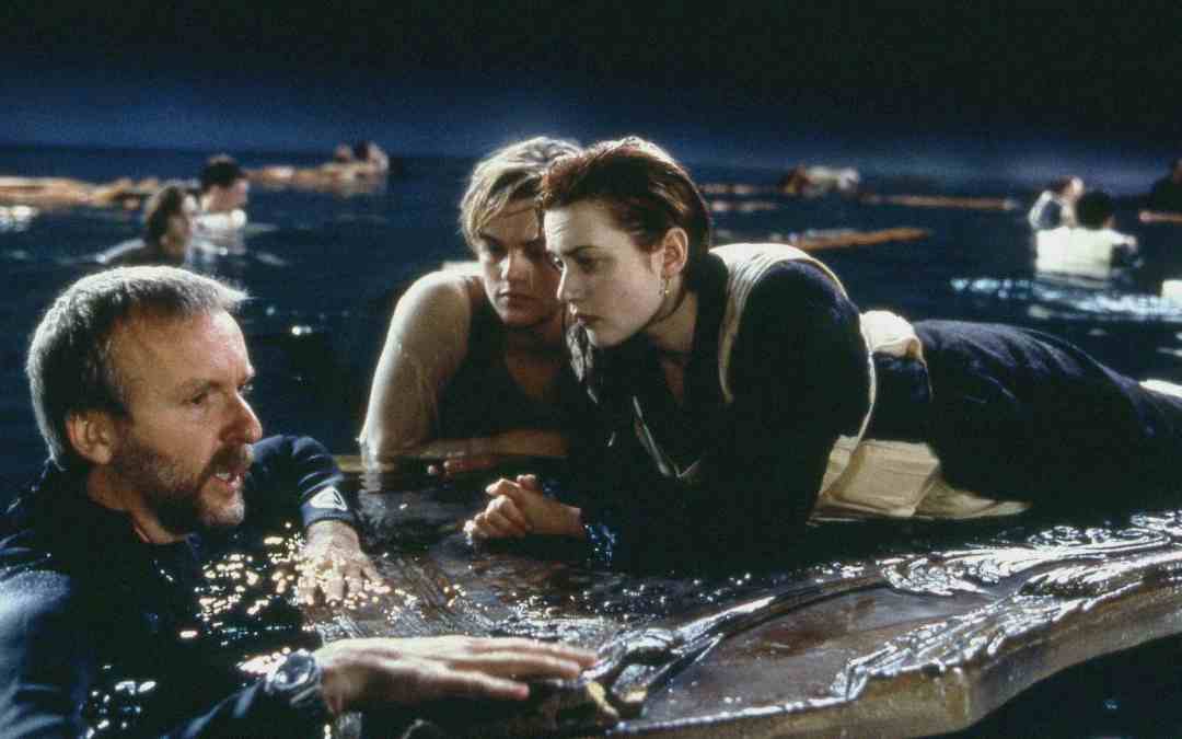 A film director talks to two actors lying on a floating wooden panel in a water tank, with people in life jackets and debris visible in the background, recreating a shipwreck scene.