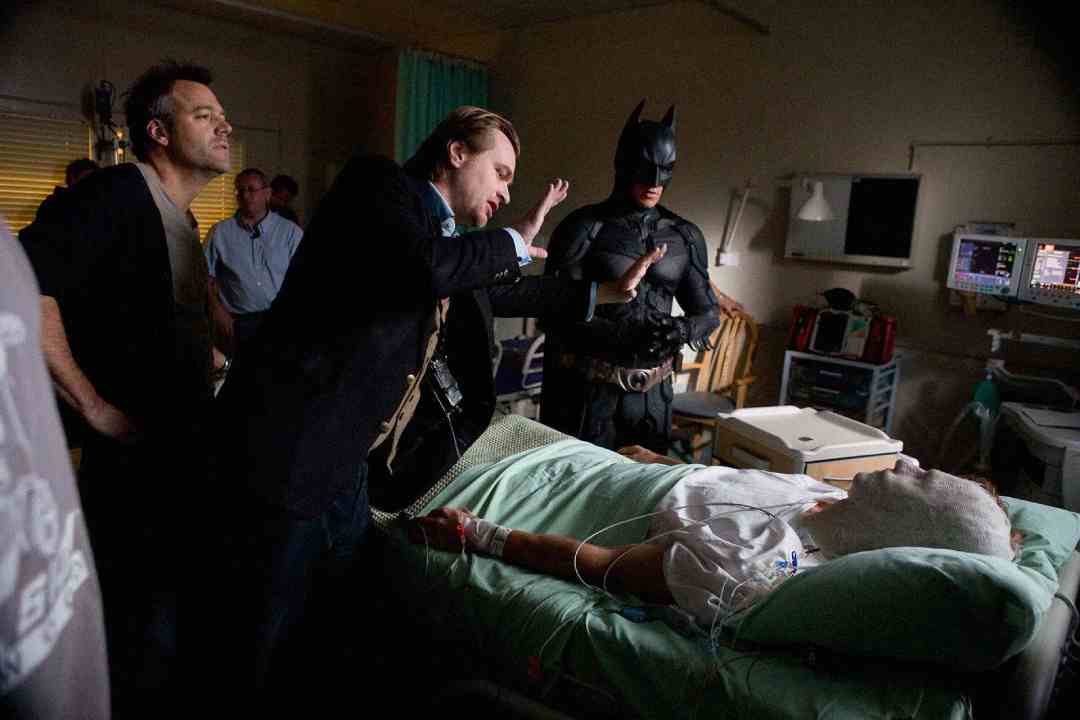 A film director gestures while talking to an actor in a Batman costume on a hospital set, where another actor lies in a hospital bed with bandages and medical equipment. Crew members and filming equipment are visible in the background.