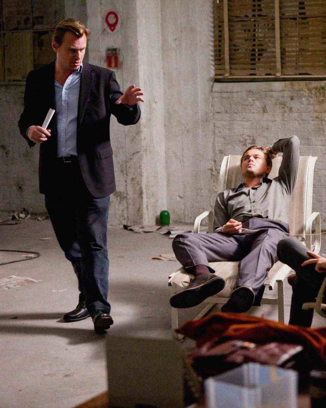 A man in a suit stands and gestures while another man lounges on a plastic chair with one arm behind his head in a dimly lit, industrial-looking room.