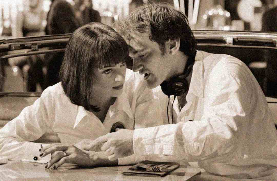 A woman with a bob haircut and a man with headphones around his neck sit closely at a table, intently looking at something as the man gestures with his hand. The image has a vintage, sepia tone.