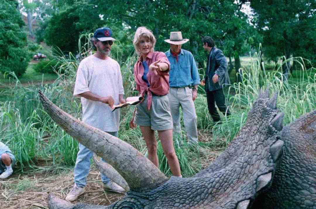 Three people stand near a large animatronic dinosaur, with lush greenery in the background. One woman points forward while a man in a white T-shirt and cap holds a script or notebook.