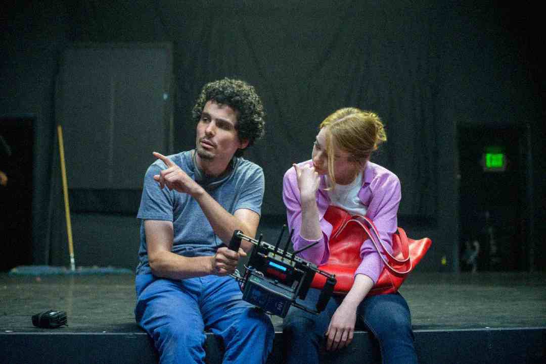 A man and a woman sit on a stage in casual clothes. The man points to the side while holding a camera rig, and the woman, holding a red bag, looks where he’s pointing. The background is dark and out of focus.