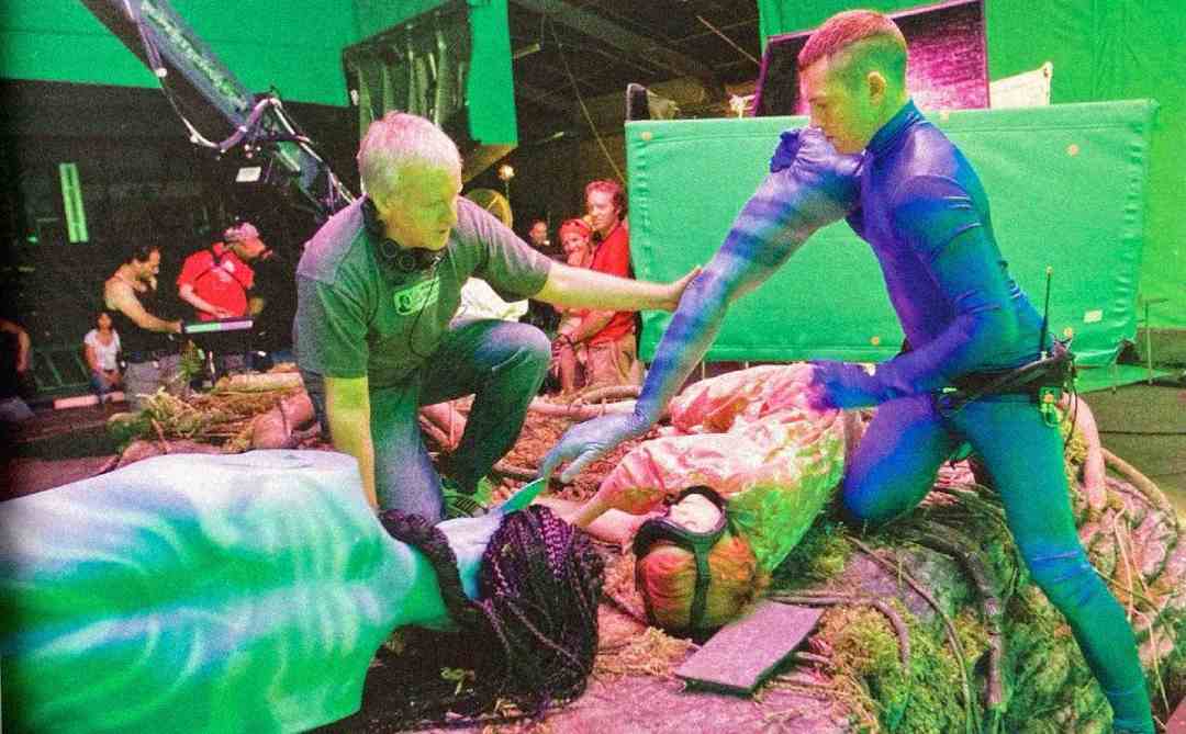 Two actors in motion capture suits and a crew member are filming a scene on a set with green screens and props. One actor is lying down while the other, in blue, reaches toward them. Production equipment is visible in the background.