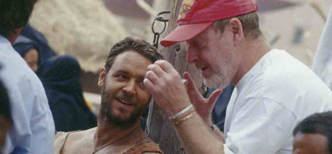 Two men are talking on a movie set. One is wearing a costume with a leather strap and smiling, while the other, in a red cap and white shirt, gestures with his hands. People and set pieces are visible in the background.