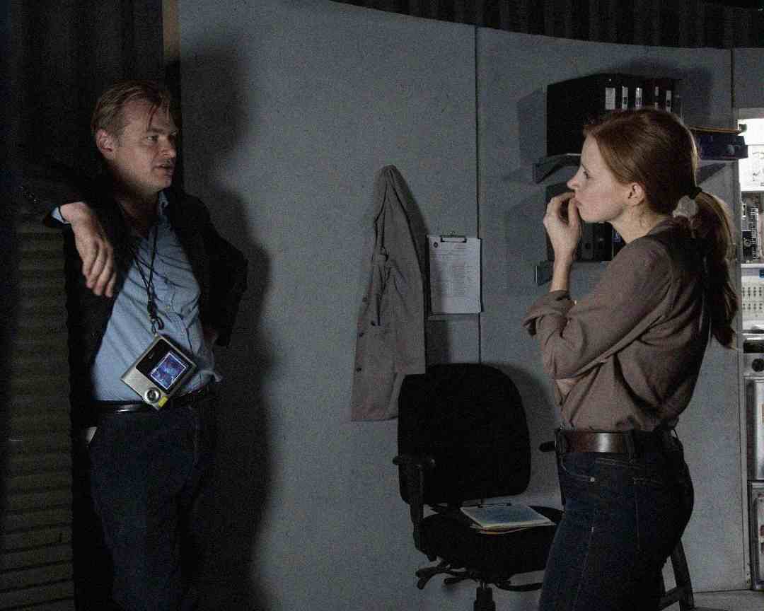 A man leans against a wall talking to a woman who is standing and eating. The room has a chair, jacket on a hook, clipboard, and office equipment, creating an informal, workplace setting.