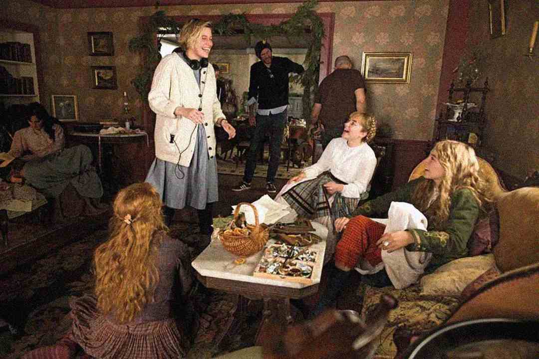 A director smiles and talks to three actresses in period costumes on a cozy, vintage living room film set, as crew members work in the background. The scene feels lively and collaborative.