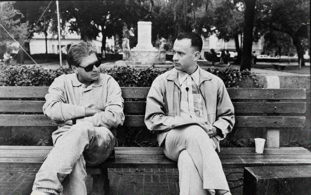 Two men sit on a park bench having a conversation. One wears sunglasses and casual clothes with arms folded, while the other is dressed more formally and sits upright. Trees and a monument are visible in the background.