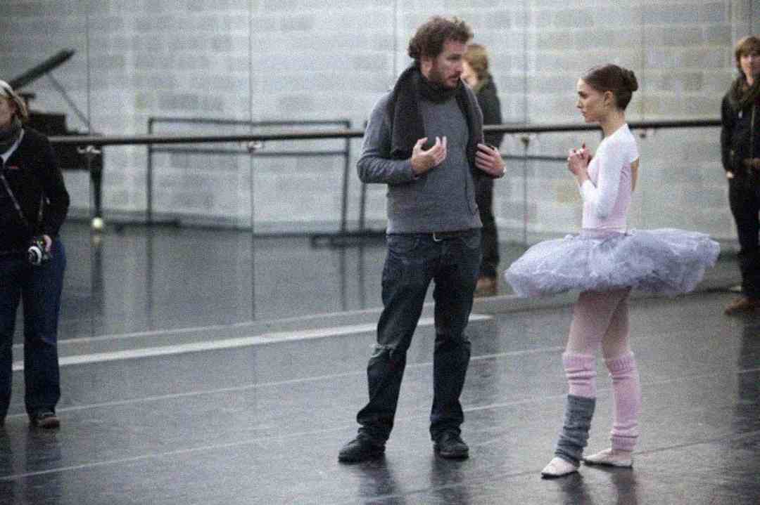 A man in casual clothes talks to a ballerina in a grey tutu and pink leg warmers in a dance studio, with mirrors and other people in the background.