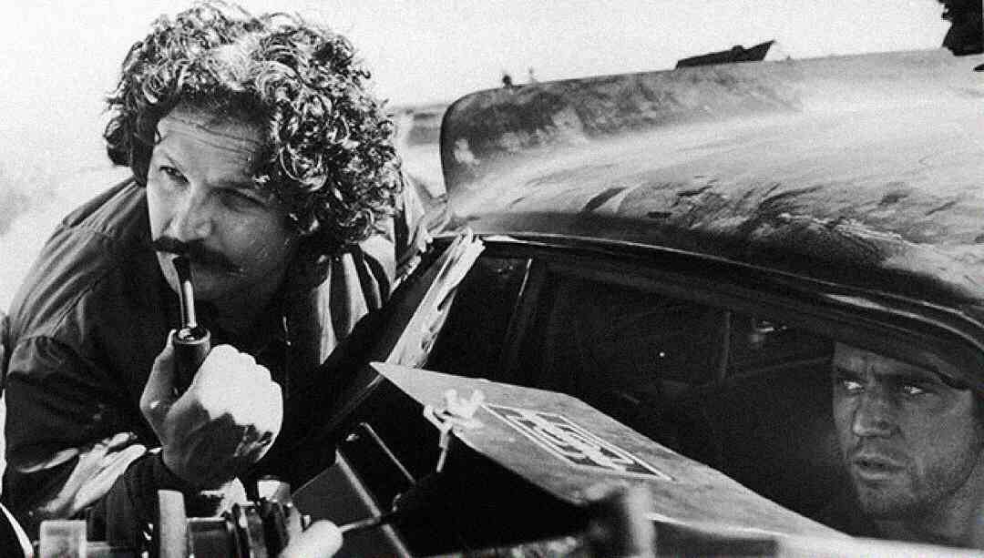 A man with curly hair and a mustache smokes a pipe while leaning on a battered car. Another man sits inside the car, looking out with a tense expression. The scene is in black and white.