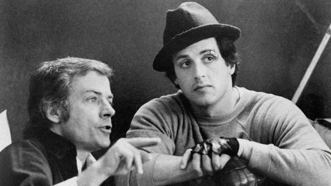 Two men speak together; one gestures while talking, and the other, wearing a hat, gloves, and a sweatshirt, looks attentively ahead. The image is black and white and has a candid, behind-the-scenes feel.