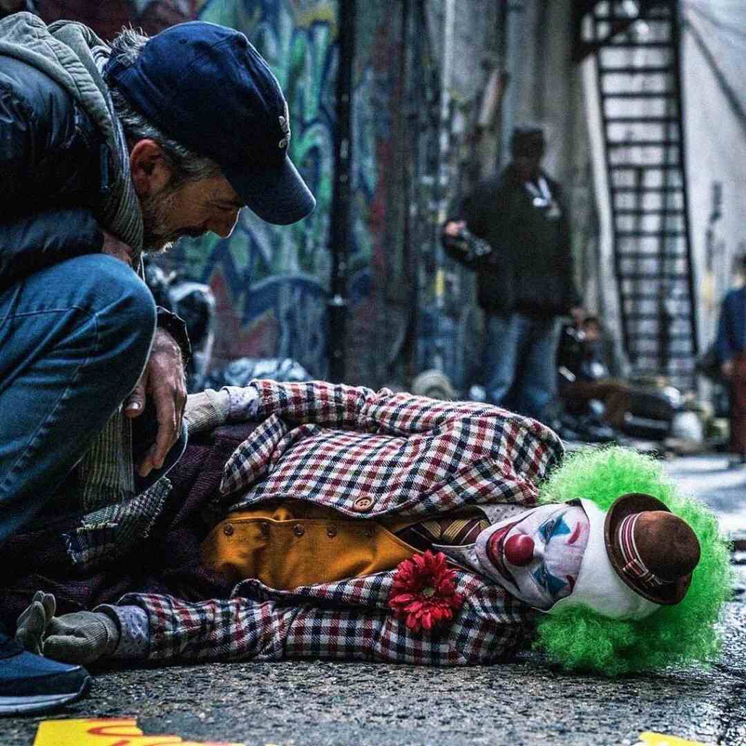 A man kneels beside a person dressed as a clown with a green wig, red nose, and checkered jacket, lying on the ground in a graffiti-covered alley. Other people and a metal staircase are visible in the background.