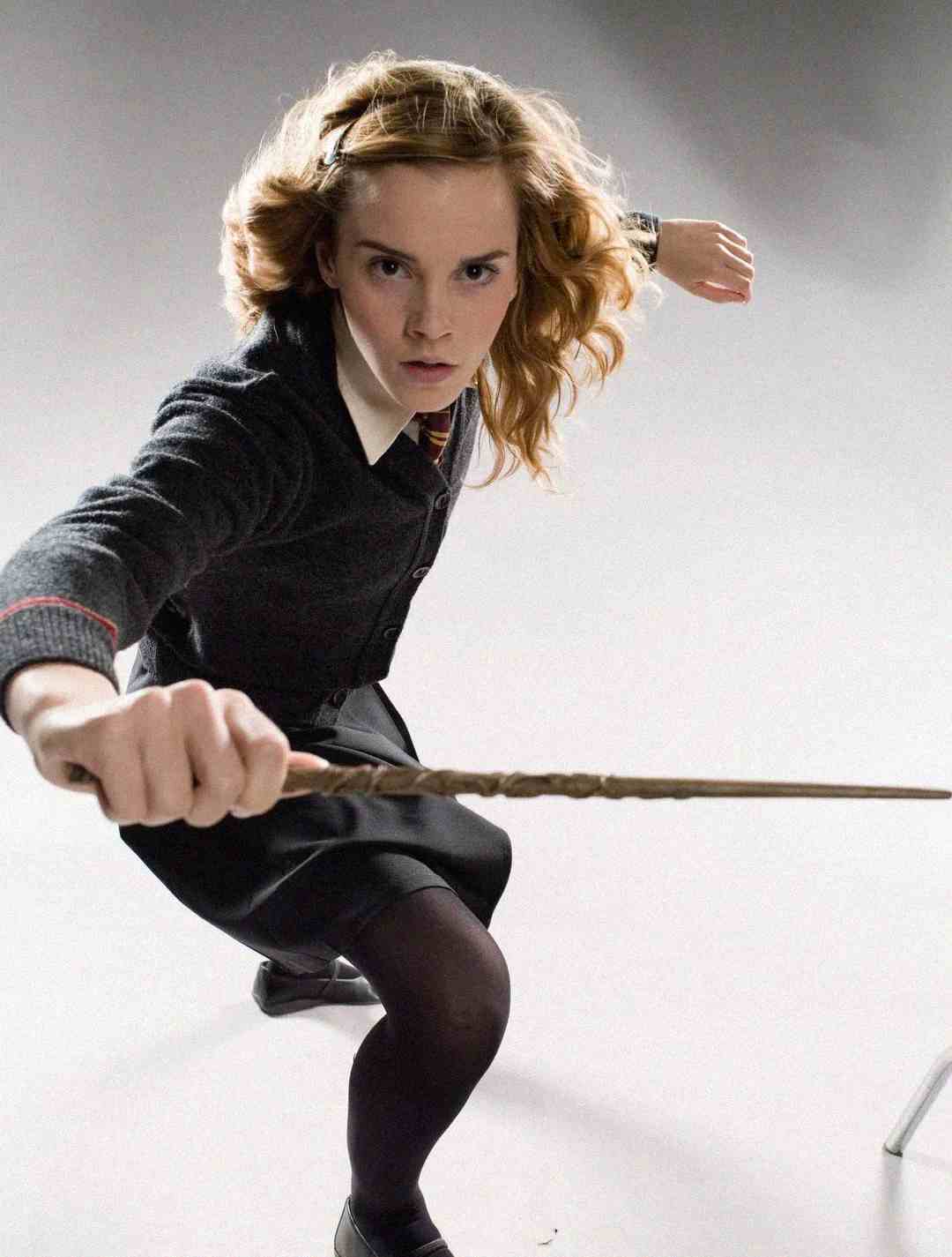 A young woman in a school uniform crouches while holding a wand out in front of her, as if casting a spell. She has wavy brown hair and a determined expression, with a white background behind her.