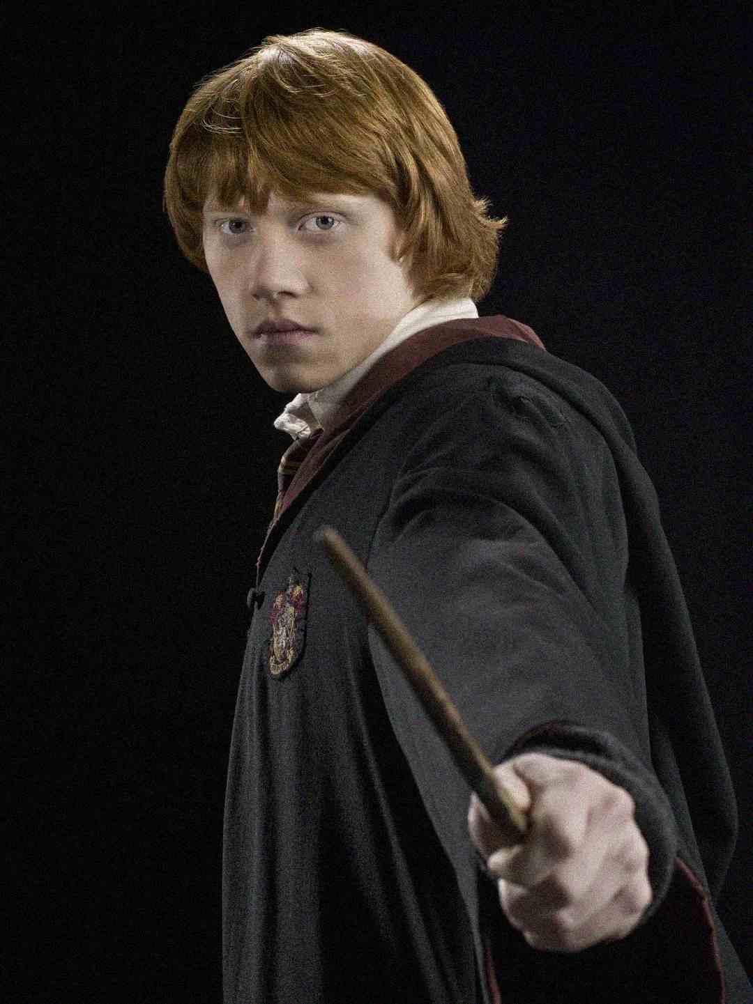 A young person with red hair, wearing a dark cloak with a crest, holds a wand pointed outward, standing against a dark background.
